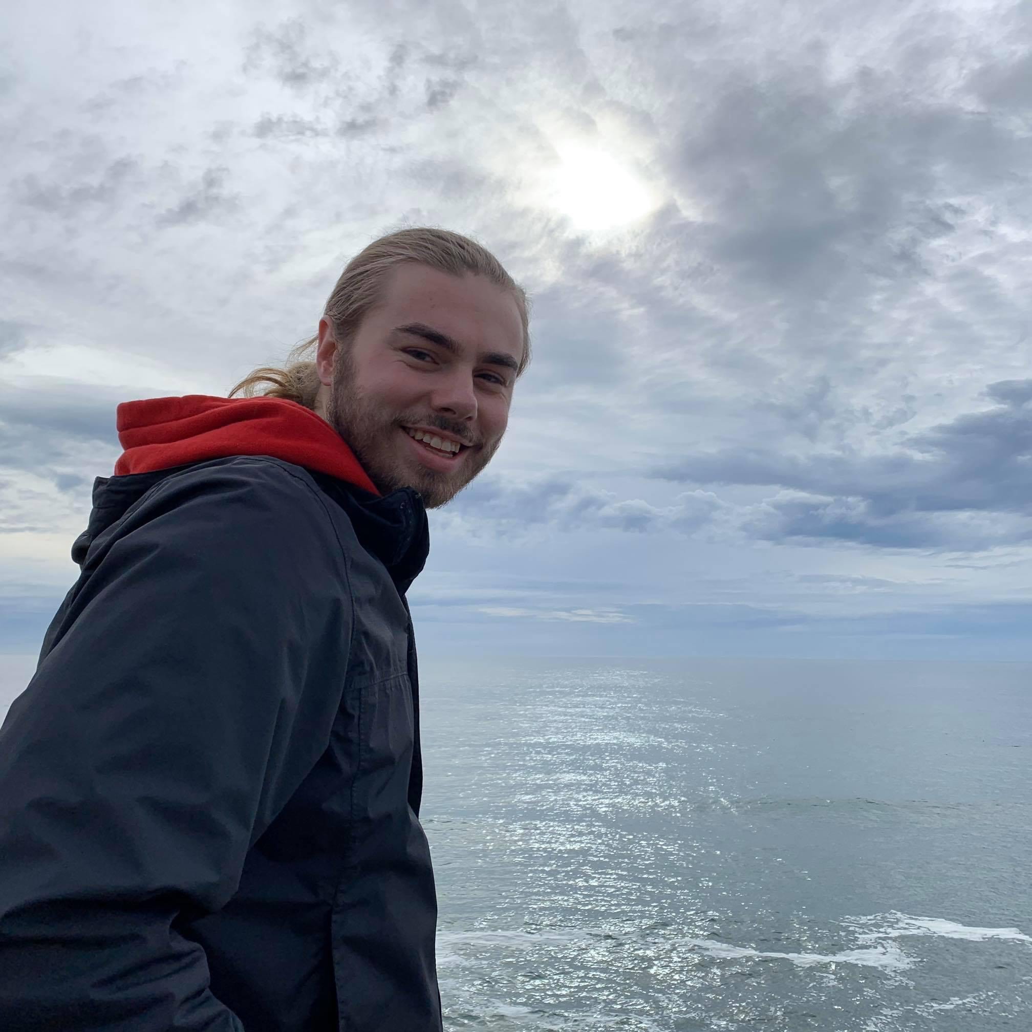 Vernon, smiling, standing in front of the ocean on a cloudy day